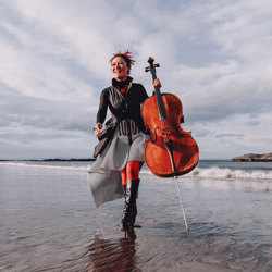 Woman in grey long dress and black short jacket running along the beach holding a cello in her left hand. Landscape shows grey skies and sea shore just behind her.